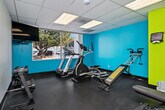 This fitness center is a thoughtfully designed exercise space that makes efficient use of its single-floor layout. The room is immediately distinguished by its bold, energizing color scheme — vivid teal and lime green accent walls create a motivating atmosphere against a neutral gray rubber flooring and a drop-panel ceiling with integrated fluorescent lighting.

A large window along one wall allows natural light to fill the workout area while providing an outdoor view, contributing to an open and airy feel. The cardio zone features a treadmill and an elliptical machine positioned near the window, making for an engaging workout experience. A cable pull station and adjustable weight bench offer options for strength training, while a rack of dumbbells along one wall supports a range of free-weight exercises.

Full-length mirrors line one wall, visually expanding the space and allowing users to monitor their form during exercise. A wall-mounted flat-screen television adds an entertainment element to the cardio area. Practical amenities include a paper towel dispenser, a water cooler, and a waste bin. Access to the fitness center is via a clean, well-lit hallway with wood-look plank flooring, providing a smooth transition into the workout space.