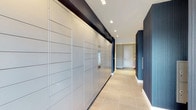 This single-floor corridor is designed with both functionality and modern aesthetics in mind. The most prominent feature is a full-length wall of smart package lockers, offering residents a secure and convenient system for receiving deliveries. The lockers are finished in a clean, light grey tone and span the entire length of one side of the hallway, providing substantial storage capacity.

The corridor is finished with large-format, light-toned tile flooring that adds a sense of cleanliness and openness to the space. The walls feature a striking combination of dark textured paneling and muted blue-grey tones, creating a contemporary and polished ambiance. Recessed square lighting runs along the ceiling, providing consistent, even illumination throughout the hallway.

At the far end of the corridor, a white door provides access to an adjoining space, while the clean architectural lines guide the eye naturally down the passage. The contrast between the dark upper wall accents and lighter lower finishes gives the space a refined, layered visual depth. Overall, the corridor presents a well-designed, practical environment with a distinctly upscale, modern character.