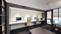 This single-floor business center is thoughtfully designed to accommodate both individual productivity and group collaboration. The space is divided into two distinct functional zones seamlessly connected by a cohesive contemporary aesthetic.

The workstation area features a long, floating white countertop running along the wall, housing multiple iMac computers separated by privacy dividers. Ergonomic cream-upholstered task chairs accompany each station, and a multifunction printer is conveniently positioned at one end. Recessed accent lighting above the workstation wall provides focused illumination, while the dark gray accent band along the upper wall adds a refined architectural contrast.

The meeting area offers a dark wood rectangular table with upholstered side chairs, suitable for small group gatherings or informal discussions. A striking sputnik-style chandelier serves as an elegant focal point overhead. Floor-to-ceiling black-framed glass partition walls divide sections of the space while maintaining visual openness and allowing light to flow through.

Underfoot, a bold dark carpet with sweeping white linear patterns ties both zones together, reinforcing the space's modern character. The overall ambiance is polished, professional, and well-suited for business productivity.