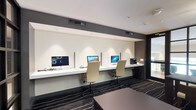 This single-floor business center is thoughtfully designed to accommodate both individual productivity and group collaboration. The space is divided into two distinct functional zones seamlessly connected by a cohesive contemporary aesthetic.

The workstation area features a long, floating white countertop running along the wall, housing multiple iMac computers separated by privacy dividers. Ergonomic cream-upholstered task chairs accompany each station, and a multifunction printer is conveniently positioned at one end. Recessed accent lighting above the workstation wall provides focused illumination, while the dark gray accent band along the upper wall adds a refined architectural contrast.

The meeting area offers a dark wood rectangular table with upholstered side chairs, suitable for small group gatherings or informal discussions. A striking sputnik-style chandelier serves as an elegant focal point overhead. Floor-to-ceiling black-framed glass partition walls divide sections of the space while maintaining visual openness and allowing light to flow through.

Underfoot, a bold dark carpet with sweeping white linear patterns ties both zones together, reinforcing the space's modern character. The overall ambiance is polished, professional, and well-suited for business productivity.