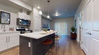 This single-floor residence offers a thoughtfully designed open-concept layout that seamlessly connects the kitchen, dining, and living areas. The kitchen is a standout feature, equipped with stainless steel appliances including a refrigerator, range, microwave, and dishwasher. White shaker-style cabinetry provides ample storage, while the contrasting dark-toned island with a white quartz countertop doubles as a breakfast bar with seating for two. Pendant lighting above the island and recessed ceiling lights enhance the functional yet stylish atmosphere. Wood-style flooring runs throughout the main living areas, adding warmth and continuity.

The living room is bright and welcoming, anchored by a large sliding glass door fitted with vertical blinds that allow natural light to filter through. The bedroom features plush carpeting, dual windows dressed with horizontal blinds, and generous natural light. A walk-in closet off the bedroom offers substantial storage space with built-in hanging rails. The full bathroom is clean and bright, featuring white cabinetry with quartz countertops, a combination tub and shower, and wood-style flooring. Neutral gray tones throughout the space create a cohesive, contemporary aesthetic.