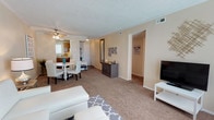 This single-story residence offers a well-connected layout that integrates the living, dining, and kitchen areas into a cohesive, open flow. The living room is carpeted and generously sized, accommodating a seating area anchored by a patterned area rug alongside a dedicated media wall with a flat-screen TV and storage unit. A ceiling fan overhead provides both lighting and ventilation, contributing to the room's functional comfort.

Adjacent to the living space, a dining area features a round white table with matching chairs, positioned near a window that allows natural light to filter in. This seating area transitions directly into the kitchen, which is visible in the background and features warm wood cabinetry, countertop workspace, and a window above the sink.

The space is finished with neutral beige walls and a textured ceiling throughout, lending a consistent and understated aesthetic. A hallway extends from the main living area, suggesting access to additional private rooms beyond. Decorative accents, including wall art and a mirrored console, add character to the otherwise clean and practical interior.