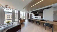 This single-floor space exemplifies contemporary design through a carefully curated open-plan layout. Floor-to-ceiling windows line the perimeter, flooding the interior with natural light while framing sweeping elevated city views. Light-toned hardwood flooring extends throughout, providing a seamless visual connection between the distinct functional zones.

The main living area is anchored by a striking marble fireplace with a clean linear profile, set against a textured slatted wood accent wall. Overhead, a sculptural wood slat ceiling detail adds warmth and architectural depth, complemented by integrated linear LED lighting. A generous lounge zone features plush neutral and gray upholstered seating accented with blue tonal cushions, paired with a long wood bar counter lined with sleek dining chairs.

A secondary seating area near the expansive windows offers an intimate setting with contemporary sofas and mid-century-inspired accent chairs. A sculptural multi-arm chandelier serves as a focal point in this zone. A compact utility area includes a clean white countertop with an undermount sink, warm wood cabinetry, stainless steel appliances, and a vibrant abstract artwork, providing practical functionality without sacrificing aesthetic cohesion. The overall ambiance is refined, airy, and modern.