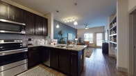 This single-floor residence offers a thoughtfully designed open-concept layout that seamlessly connects the kitchen, dining, and living areas. The kitchen is a standout feature, outfitted with dark espresso cabinetry, granite countertops, a tile backsplash, stainless steel appliances including a French door refrigerator and range, and pendant lighting over a peninsula with bar seating. Wood-style flooring runs throughout the main living spaces, adding warmth and continuity.

The living room is anchored by a striking deep teal accent wall, a ceiling fan, and large windows that allow generous natural light to fill the room. A door leads to an outdoor area, extending the living space beyond the interior.

The bedroom continues the teal accent wall motif, creating a cohesive design aesthetic. It features plush carpeting, multiple windows that bring in abundant natural light, and direct access to a private en-suite bathroom equipped with dark cabinetry and quality finishes. A dedicated dressing or hallway area with additional storage connects the bedroom to the bath, adding functional convenience.

Throughout, the design is unified by a palette of deep teal, warm neutrals, and rich dark wood tones, resulting in a modern, stylish, and comfortable living environment.