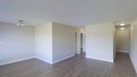 This single-floor space presents a clean and understated interior design, characterized by neutral beige and white tones that create a bright, airy atmosphere. The main living area features durable hardwood-style flooring in a warm medium-toned wood finish, which extends throughout the space and provides a cohesive visual flow. Smooth white ceilings at a standard height complement the simple, unadorned walls finished in a soft greige tone, contributing to the overall sense of calm and versatility.

The open layout of the main area is defined by a partial wall that subtly separates spaces while maintaining an open feel. White baseboards provide clean contrast against both the walls and flooring. A hallway visible in the background leads to additional rooms, including what appears to be a closet or utility space behind one door and a further room beyond another. Electrical outlets are evenly distributed along the walls, and a ceiling-mounted light fixture is visible near the hallway entry. The space is move-in ready with a neutral palette suited to a wide range of furnishing styles.