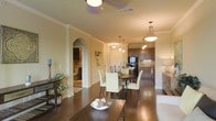This single-story home showcases an open-concept design that seamlessly connects the living, dining, and kitchen areas. The dining space is anchored by a dark wood table surrounded by cream upholstered chairs, illuminated by a pendant light fixture that adds a warm, inviting glow. Rich hardwood flooring flows throughout, complementing the neutral warm tones of the walls and crown molding that frames each room with classic elegance.

The kitchen, visible from the dining area, features dark wood cabinetry, stainless steel appliances including a refrigerator, and a functional layout that offers ample counter space. Pendant lighting above the kitchen area enhances both practicality and ambiance.

A graceful arched doorway provides a view into an adjoining room, adding architectural character and a sense of depth to the space. Crown molding details and recessed ceiling vents are consistent throughout, reflecting thoughtful construction. A ceiling fan with integrated lighting serves the main living area, ensuring comfort. Decorative touches, including potted greenery and a sideboard with a table lamp, contribute to the warm, lived-in feel of this well-appointed home.