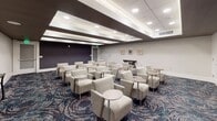 Located at 7770 Farr St in Charleston, SC, this single-floor meeting and presentation room offers a well-appointed, professionally designed space suited for seminars, screenings, or group gatherings. The room is furnished with rows of upholstered lounge chairs in a neutral gray fabric, each paired with compact round side tables, creating a comfortable yet functional seating arrangement for attendees. A large flat-panel display screen is mounted at the front of the room, making it well-equipped for presentations and media viewing. The ceiling features a distinctive coffered design with dark accent beams and recessed lighting that provides bright, even illumination throughout the space. The boldly patterned carpet in deep navy, teal, and burgundy tones adds a dynamic visual element to the room's otherwise understated color palette of soft whites and grays. One wall is clad in a textured dark tile panel, offering a striking architectural contrast. A decorative console table and framed artwork along the walls contribute an elevated, refined aesthetic. A built-in beverage station or cabinetry unit is positioned at the rear of the room for added convenience. The overall ambiance is polished and contemporary, balancing professional functionality with thoughtful interior design details.