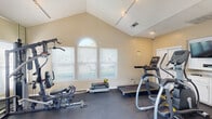 Located in Baton Rouge, Louisiana, this dedicated fitness center offers a well-appointed workout space designed to support a full range of exercise routines. The room is defined by its striking vaulted ceiling, which rises to a dramatic peak and lends the space an open, airy feel that extends well beyond its compact footprint. A combination of track lighting and recessed fixtures ensures the room is thoroughly illuminated throughout the day and evening.

Natural light flows in generously through multiple windows fitted with horizontal blinds, including a distinctive arched window that serves as a focal architectural detail. A set of French doors opens directly to an outdoor pool area, reinforcing the connection to the broader community amenities and allowing for excellent ventilation.

The fitness floor is finished with durable dark rubber flooring, providing a safe and functional surface for all types of training. Equipment includes multiple treadmills, an elliptical trainer, a stationary recumbent bike, and a multi-station cable strength machine, offering both cardiovascular and resistance training options. A full-length wall mirror on one side of the room enhances the sense of space while allowing users to monitor their form. Storage closets and a wall-mounted television round out the practical amenities of this thoughtfully outfitted community fitness center.