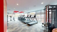 Located at 65 Brainerd Road in Boston's Allston neighborhood, this well-appointed fitness center offers a spacious and thoughtfully designed exercise environment across a single floor. The room is flooded with natural light through expansive floor-to-ceiling windows that line multiple walls, providing sweeping views of the surrounding Boston streetscape and creating an open, energizing atmosphere.

The interior design is defined by bold red accent panels on the white ceiling and striking red accent walls in the strength training area, adding a dynamic visual energy to the space. Geometric-patterned carpet tiles in neutral tones cover the floor throughout, complementing the clean, modern aesthetic.

The cardio zone features a row of treadmills and elliptical trainers, all equipped with individual digital displays, positioned directly facing the panoramic windows. A dedicated strength training section includes a comprehensive cable/functional trainer, a full dumbbell rack with weight benches, and a TRX-style suspension system, all set against the signature red accent walls and mirrored surfaces. Additional equipment includes a rowing machine, stationary bikes, stability balls, and a balance board. A hydration station with a water filtration unit is conveniently mounted on the wall. Recessed ceiling lighting supplements the abundant natural light, ensuring a bright and welcoming workout environment throughout the day.