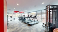 Located at 65 Brainerd Road in Boston's Allston neighborhood, this well-appointed fitness center offers a spacious and thoughtfully designed exercise environment across a single floor. The room is flooded with natural light through expansive floor-to-ceiling windows that line multiple walls, providing sweeping views of the surrounding Boston streetscape and creating an open, energizing atmosphere.

The interior design is defined by bold red accent panels on the white ceiling and striking red accent walls in the strength training area, adding a dynamic visual energy to the space. Geometric-patterned carpet tiles in neutral tones cover the floor throughout, complementing the clean, modern aesthetic.

The cardio zone features a row of treadmills and elliptical trainers, all equipped with individual digital displays, positioned directly facing the panoramic windows. A dedicated strength training section includes a comprehensive cable/functional trainer, a full dumbbell rack with weight benches, and a TRX-style suspension system, all set against the signature red accent walls and mirrored surfaces. Additional equipment includes a rowing machine, stationary bikes, stability balls, and a balance board. A hydration station with a water filtration unit is conveniently mounted on the wall. Recessed ceiling lighting supplements the abundant natural light, ensuring a bright and welcoming workout environment throughout the day.
