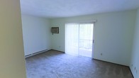 This single-floor space offers a straightforward and functional layout designed for comfortable everyday living. The main living area is finished with wall-to-wall carpet flooring, providing a soft and quiet underfoot experience. A large sliding glass door, dressed with vertical blinds, serves as the focal point of the room and fills the space with an abundance of natural light while offering direct access to an outdoor area such as a balcony or patio. A wall-mounted air conditioning unit provides climate control, complemented by baseboard heating along the perimeter walls for year-round comfort. The walls are finished in a neutral, light tone that enhances the brightness of the room and contributes to its clean, airy atmosphere. The overall design is simple and uncluttered, making the space highly adaptable to a variety of furniture arrangements and personal styles. The connection to the outdoors through the sliding glass door adds a sense of openness, bringing in views of surrounding greenery and making the living area feel more expansive than its footprint might suggest.