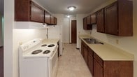 This single-floor space offers a straightforward, functional layout with well-defined living areas connected by a central corridor. The main living area is generously proportioned, featuring neutral-toned wall-to-wall carpeting, white walls, and a window air conditioning unit. A large sliding glass door provides direct access to a balcony, allowing natural light to fill the space and offering views of surrounding greenery. Baseboard heating runs throughout the carpeted rooms, ensuring consistent warmth.

The layout includes multiple carpeted rooms that function as bedrooms or flexible living spaces. One room features two windows fitted with horizontal blinds, drawing in ample natural light and views of mature trees outside. A textured ceiling is present in several rooms, adding a subtle decorative element.

The galley-style kitchen is equipped with dark wood cabinetry lining both walls, a white electric coil-burner range, a refrigerator, a dishwasher, and a double basin sink with light-toned countertops. Tile-style flooring runs through the kitchen and connects to the hallway.

The bathroom includes a full tub-shower combination with tiled surround, a toilet, a vanity with a light countertop, and tile flooring. A wood-panel door with a brass knob completes the space.