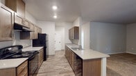 Located at 1203 NE Territorial Rd in Canby, Oregon, this single-floor residence offers a well-organized layout with a clean, modern aesthetic throughout. The galley-style kitchen is a standout feature, equipped with warm wood cabinetry, light countertops, black appliances including a range and refrigerator, and a double-basin sink. Wood-look flooring runs through the kitchen and utility areas, adding warmth and durability. The living area is generously proportioned, finished with plush wall-to-wall carpeting and neutral-toned walls that create a calm, inviting atmosphere. The bedrooms continue this theme with soft carpet underfoot and windows fitted with vertical blinds that allow natural light while maintaining privacy. Two bathrooms serve the residence — one with a walk-in shower stall and single vanity, the other with a toilet and vanity — both featuring wood-look flooring, white Craftsman-style panel doors, and matte black hardware accents. A dedicated in-unit laundry area includes both a washer and dryer with an overhead shelf for added storage. The overall design favors practicality and comfort, with consistent finishes throughout that create a cohesive and move-in-ready living environment.