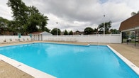 This amenity space offers residents a comprehensive recreational experience, combining outdoor leisure with indoor fitness facilities. The centerpiece of the outdoor area is a generously sized swimming pool with crystal-clear blue water, surrounded by a clean concrete deck lined with white lounge chairs — ideal for relaxing during warmer months. A white privacy fence encloses the pool area, and a small utility shed is conveniently located nearby. Mature trees in the background provide a natural backdrop.

Inside, the fitness center is thoughtfully designed with light wood-tone flooring and a neutral color palette accented by a teal feature wall. The space is well-lit by recessed lighting and natural light from multiple windows. The cardio zone features a row of modern treadmills and elliptical machines, all equipped with individual digital screens. The strength training area is stocked with a full array of Life Fitness cable machines, weight benches, free weights, and a dumbbell rack. A water cooler station is conveniently positioned near the entrance for easy access. Additional rooms off the main fitness floor provide supplementary space, finished with blue-toned carpet tile flooring and clean white walls with gray trim.