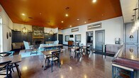 This generously sized single-floor community gathering space offers a well-appointed interior designed for socializing, relaxation, and events. The space is divided into distinct functional zones that flow seamlessly into one another across its expansive polished concrete floors.

The main lounge area serves as a focal point, anchored by a full brick fireplace and framed by dark navy beadboard wainscoting. Warm orange-painted ceilings with globe pendant chandeliers add a bold, inviting character to this zone, which is furnished with a plush sectional sofa, accent chairs, and casual dining tables with cross-back chairs. A wall-mounted flat-screen TV enhances the space for entertainment.

Adjacent to the lounge, a bar and counter area features a full-length service counter with bar stools, gray paneled cabinetry, pendant lighting, and upper storage — ideal for community events or casual gatherings. A separate, brighter room benefits from large multi-pane windows that flood the space with natural light and offer views of an outdoor pool and courtyard. This room includes additional seating, a shuffleboard table, and a projection screen for presentations or screenings.

Throughout, the design balances rustic and contemporary elements, with decorative wall art, vintage-style clocks, and layered lighting creating a warm, welcoming ambiance suitable for a variety of uses.