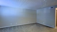 This single-story space presents a clean, move-in-ready interior highlighted by a well-proportioned room visible in the available imagery. The room features wall-to-wall carpeting in a neutral beige tone, providing a soft and comfortable underfoot experience. The walls are finished in a light gray-beige palette, offering a versatile backdrop suited to a variety of furnishing styles and décor preferences. A textured popcorn ceiling adds dimension overhead, complementing the overall understated aesthetic of the space. Natural light enters through windows fitted with horizontal blinds, allowing for adjustable light control while contributing to a bright and airy atmosphere. Standard electrical outlets are positioned along the baseboards, indicating practical utility throughout. The white baseboard trim provides clean definition between the walls and flooring, adding a crisp finishing detail. The room's open layout offers flexibility for multiple furniture configurations. Overall, the space maintains a neutral, functional character with a simple and tidy interior finish that serves as a solid foundation for personalization.