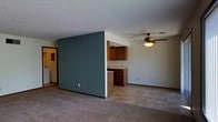 This single-floor space is defined by an open-concept layout that connects the living area and kitchen through a thoughtful half-wall partition. The partial wall, painted in a muted teal-green tone, serves as a visual divider while maintaining an open and airy feel between the two spaces. On the kitchen side, warm wood-toned cabinetry is mounted above a countertop with a built-in lower cabinet unit, providing functional storage and workspace. The kitchen area transitions into tile-style flooring, while the living space features wall-to-wall carpeting in a neutral beige tone, adding warmth underfoot. A ceiling fan with a built-in light fixture is centrally positioned in the living area, offering both ventilation and ambient lighting. The ceilings feature a textured finish throughout, and the walls are painted in a clean, neutral white that complements the teal accent wall. A doorway visible on the far left suggests additional rooms or storage beyond the main living space. Overall, the layout is practical and straightforward, designed to maximize usable space within a compact, single-level footprint.