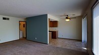 This single-floor space is defined by an open-concept layout that connects the living area and kitchen through a thoughtful half-wall partition. The partial wall, painted in a muted teal-green tone, serves as a visual divider while maintaining an open and airy feel between the two spaces. On the kitchen side, warm wood-toned cabinetry is mounted above a countertop with a built-in lower cabinet unit, providing functional storage and workspace. The kitchen area transitions into tile-style flooring, while the living space features wall-to-wall carpeting in a neutral beige tone, adding warmth underfoot. A ceiling fan with a built-in light fixture is centrally positioned in the living area, offering both ventilation and ambient lighting. The ceilings feature a textured finish throughout, and the walls are painted in a clean, neutral white that complements the teal accent wall. A doorway visible on the far left suggests additional rooms or storage beyond the main living space. Overall, the layout is practical and straightforward, designed to maximize usable space within a compact, single-level footprint.