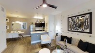 This thoughtfully designed single-floor space seamlessly blends modern aesthetics with functional living. The open-concept layout connects the living area and kitchen, creating an inviting, airy atmosphere ideal for both relaxing and entertaining.

The living area is anchored by a plush, neutral-toned sofa accented with dark throw pillows, a textured area rug, and a sleek glass coffee table. A striking piece of wall art and decorative chrome accents add a contemporary flair, while a stylish floor lamp provides warm, ambient lighting. A modern three-blade ceiling fan with an integrated light fixture enhances airflow and adds a bold design element overhead.

The kitchen features dark cabinetry contrasted by light countertops and a grey breakfast bar peninsula, which doubles as a casual dining or prep space. Stainless steel appliances, including a full-size refrigerator, complement the modern design, while industrial-style pendant lights hang above the peninsula, adding character and focused illumination.

Wood-look flooring runs throughout the space, offering a warm, cohesive feel that ties each area together. A glimpse of an adjacent dining area with a mirror and natural light further enhances the sense of openness. Overall, the space exudes a clean, contemporary elegance.