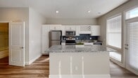 This single-floor residence offers a thoughtfully designed layout that balances functionality with modern style. The kitchen serves as a central highlight, showcasing crisp white cabinetry, granite countertops, and a full suite of stainless steel appliances including a refrigerator, electric range, and over-the-range microwave. A tiled backsplash in cool blue-gray tones adds a contemporary accent, while a built-in wine rack provides a charming finishing touch. The kitchen opens to a peninsula counter that seamlessly connects to the living area, making it ideal for both daily use and entertaining. The living space is clean and inviting, featuring warm wood-look flooring, neutral wall tones, and a ceiling fan that enhances comfort. A window brings in natural light, contributing to the room's airy feel. The bathroom is well-equipped with a combination tub and shower enclosed in gold-framed glass doors and accented with marble-style tile. A tall white storage cabinet, granite-topped vanity, and wood-look flooring complete the space with a polished, cohesive look. Throughout the residence, consistent use of wood-look flooring, neutral tones, and modern finishes creates a unified and contemporary aesthetic.