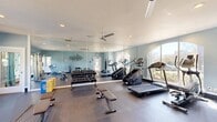 Located in Columbus, Georgia, this fitness center offers a thoughtfully designed exercise space built to support a comprehensive workout experience. The room features light blue walls accented by a bold motivational "WORKOUT" typographic wall mural, creating an energetic and inspiring atmosphere throughout. Large arched and rectangular windows line the walls, flooding the space with natural light and offering views of the surrounding outdoor area, including what appears to be a pool courtyard. A full-length wall mirror expands the sense of space visually while aiding exercisers in monitoring their form. Ceiling fans provide comfortable airflow during intense sessions. The flooring transitions between rubber matting and hard surface zones, catering to a variety of workout types. Equipment is well-distributed and includes treadmills, a stair climber, a rowing machine, and a dual cable functional trainer for strength training. A tiered dumbbell rack stocked with a full range of free weights sits alongside flat workout benches. Medicine balls and stability balls are neatly stored on dedicated racks, and a water cooler ensures easy hydration. Recessed lighting supplements the abundant natural light for consistent illumination at any hour. The space is efficiently organized to maximize usability without feeling crowded.