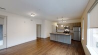 This contemporary single-floor residence showcases a thoughtfully designed open-concept layout that seamlessly connects the living and kitchen areas. Rich hardwood flooring extends throughout the main living space, lending warmth and continuity to the interior. The kitchen is a standout feature, centered around a large island with a built-in sink and dark countertops, offering both functional workspace and casual seating potential. Taupe-toned cabinetry lines the kitchen walls, complemented by a classic tile backsplash and a full suite of stainless steel appliances, including a refrigerator, stove, and over-the-range microwave. Pendant lighting hangs above the island, while additional track lighting illuminates the kitchen and dining area, creating a layered and inviting ambiance. The living area is bright and open, with clean white walls and ample room for furnishing. A hallway visible from the main space leads to additional rooms, suggesting a well-organized and functional floor plan. The overall aesthetic is clean, modern, and move-in ready, with contemporary finishes that strike a balance between style and practicality.