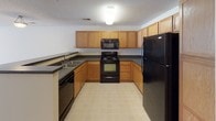 This single-floor unit is thoughtfully laid out across a practical, open floor plan. The kitchen is a functional U-shaped space outfitted with warm oak cabinetry, dark countertops, and a full suite of black appliances, including a refrigerator, electric range, built-in microwave, and dishwasher. Light tile flooring runs throughout the kitchen and extends into the hallway, providing an easy-to-maintain surface in high-traffic areas. A breakfast bar with an overhanging countertop connects the kitchen to the adjoining living area, which features plush wall-to-wall carpeting, a ceiling fan with integrated lighting, and a textured ceiling. The living space flows naturally toward the entry corridor, which includes a built-in storage closet. The bedrooms are carpeted and bathed in natural light through sizable windows fitted with horizontal blinds. The bathroom is generously proportioned, featuring a combination soaking tub and shower, a dedicated towel bar, a standard toilet, and a separate vanity area with warm oak cabinetry and a large mirror. Light tile flooring, neutral wall tones, and consistent finishes throughout lend the space a cohesive, understated aesthetic.