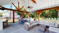 Located at 511 Cobblestone Dr in Wilmington, NC, this beautifully designed outdoor pavilion offers a sophisticated and functional open-air gathering space. The structure is defined by a striking gabled roof supported by warm, rich-toned timber-frame posts and trusses, with a crisp white paneled ceiling that reflects natural light and lends an airy, expansive feel throughout. Ceiling fans are strategically positioned to ensure comfort in the coastal Carolina climate, while string lights add a welcoming ambiance.

The pavilion is organized into distinct functional zones. A built-in outdoor kitchen anchors one end, equipped with a stainless steel gas grill and ample counter space finished in a light-toned surface, making it ideal for entertaining. Adjacent to the kitchen, a sizable dining table with ample seating provides a dedicated space for outdoor meals. The lounge area is generously appointed with oversized wicker sectional sofas and coordinating ottomans, dressed in neutral cushions with teal accent pillows, arranged around a coffee table for relaxed conversation.

The aggregate concrete flooring extends throughout the covered area and beyond, providing a durable and polished foundation. Open on all sides, the pavilion offers unobstructed views of the surrounding green lawn and mature trees, creating a seamless connection with the outdoors.