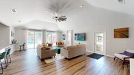 Located at 503 Bertram Ct in Augusta, GA, this single-story clubhouse offers a well-designed, open-concept layout that blends functionality with modern style. The space is defined by soaring vaulted ceilings that lend an expansive, airy feel throughout, complemented by durable wood-look flooring that runs seamlessly from room to room.

The combined living and dining area serves as the heart of the clubhouse, bathed in natural light from large sliding glass doors and multiple windows that frame views of the outdoor pool area. The living space features a dramatic windmill-style ceiling fan as a standout design element, along with comfortable seating arrangements and a wall-mounted flat-screen TV. Two dining tables with mid-century modern chairs provide ample seating for gatherings, while a built-in workstation along one wall adds versatility to the space.

The kitchen is clean and contemporary, outfitted with white shaker cabinetry, granite countertops, stainless steel appliances, and a dedicated beverage cooler. A pass-through window connects the kitchen to the main living area, maintaining the open and social atmosphere of the clubhouse.

A hallway with a glass-panel entry door provides a welcoming transition into the space, enhanced by vaulted ceilings that carry through from the main area. The overall ambiance is bright, modern, and inviting — ideal for community gatherings and events.
