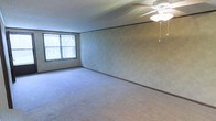 This single-floor space offers a clean, functional layout designed for comfortable everyday living. The main living area is generously proportioned, providing ample room for a variety of furniture arrangements. Soft, neutral-toned wall-to-wall carpeting covers the floor throughout, lending a warm and cohesive feel to the interior. The walls feature a subtle textured wallcovering accented by a classic chair rail molding, adding a touch of traditional character to the space. A ceiling fan with a built-in light fixture is centrally positioned overhead, offering both ambient lighting and air circulation year-round. A large window fitted with vertical blinds allows natural light to fill the room, creating a bright and airy atmosphere while maintaining privacy. The ceiling displays a textured finish consistent with the overall design of the space. The neutral color palette throughout — from the flooring to the walls — provides a versatile backdrop that can easily accommodate a range of interior styles. Overall, the space presents a straightforward, well-maintained environment that is move-in ready and adaptable to a variety of living needs.