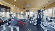 This fitness center offers a thoughtfully designed single-floor layout that accommodates a wide variety of workout routines. The space is finished with warm, neutral-toned walls accented by crown molding, recessed lighting, and a ceiling fan that helps maintain comfortable airflow throughout. Durable rubber flooring covers the entire workout area, providing both safety and support underfoot.

The strength training section is well-appointed, featuring a multi-station cable machine, a power tower for bodyweight exercises, an adjustable weight bench, and a full rack of dumbbells in varying weights. A wall-mounted flat-screen television and large mirrors enhance the environment, making the space feel even more expansive while allowing users to monitor their form.

A dedicated cardio zone is positioned near a bay of large windows, housing multiple treadmills and additional cardio equipment, including a stationary bike. The generous window coverage throughout the facility floods the interior with natural light and provides pleasant outdoor views of the surrounding greenery.

Multiple entry and exit points, including doors leading to the exterior, ensure easy accessibility. The overall ambiance is bright, open, and functional, making this fitness center a practical and inviting space for a full range of exercise needs.