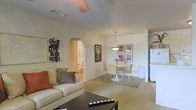 This single-floor residence offers a thoughtfully designed open-concept layout that seamlessly connects the living and dining areas, creating a cohesive and inviting atmosphere throughout. The living room is anchored by a plush, neutral-toned sofa accented with colorful throw pillows, complemented by a sleek coffee table and a decorative wall-mounted art piece that adds a contemporary touch. A ceiling fan overhead ensures year-round comfort in the space.

A graceful arched doorway serves as a standout architectural feature, lending a classic, elegant character to the interior while providing a smooth transition to adjoining areas, including a hallway that leads to additional rooms. The dining area sits adjacent to the living space and is centered around a round dining table with wicker-style chairs, set atop a natural area rug. A modern pendant light hangs above, casting warm illumination over the table, while a set of framed artwork adorns the wall, adding color and visual interest.

The neutral, light-toned walls and carpeted flooring throughout contribute to a bright, airy ambiance. A glimpse of the kitchen is visible, featuring white appliances that maintain the clean, cohesive aesthetic of the home. Overall, this residence offers a comfortable and functional living environment with charming architectural details.