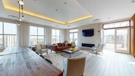 Located at 695 Atlantic St in Stamford, Connecticut, this single-floor amenity lounge is a thoughtfully designed communal space that blends modern aesthetics with warm, inviting materials. The layout seamlessly integrates a lounge area, dining zone, and kitchenette into one cohesive, open-plan environment.

The lounge area is anchored by a rich leather sofa and complementary seating arranged around a rustic wood coffee table atop a patterned area rug. A sleek linear fireplace and a wall-mounted flat-screen TV serve as focal points, while a coffered ceiling with warm ambient uplighting adds architectural depth and a refined atmosphere. Large floor-to-ceiling windows flood the space with natural light and offer expansive views of the Stamford skyline.

Separating the lounge from the dining area is a striking floor-to-ceiling slatted wood partition wall, adding warmth, texture, and a sense of artful division without fully closing off the space. The dining area features a solid wood communal table with bench seating, pendant lighting overhead, and a decorative tiled accent wall behind a built-in kitchenette with wood cabinetry. Light wood-toned flooring flows throughout, tying all areas together. Access to an outdoor terrace further enhances the space's connection to natural light and open-air city views.