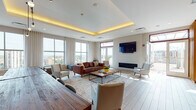 Located at 695 Atlantic St in Stamford, Connecticut, this single-floor amenity lounge is a thoughtfully designed communal space that blends modern aesthetics with warm, inviting materials. The layout seamlessly integrates a lounge area, dining zone, and kitchenette into one cohesive, open-plan environment.

The lounge area is anchored by a rich leather sofa and complementary seating arranged around a rustic wood coffee table atop a patterned area rug. A sleek linear fireplace and a wall-mounted flat-screen TV serve as focal points, while a coffered ceiling with warm ambient uplighting adds architectural depth and a refined atmosphere. Large floor-to-ceiling windows flood the space with natural light and offer expansive views of the Stamford skyline.

Separating the lounge from the dining area is a striking floor-to-ceiling slatted wood partition wall, adding warmth, texture, and a sense of artful division without fully closing off the space. The dining area features a solid wood communal table with bench seating, pendant lighting overhead, and a decorative tiled accent wall behind a built-in kitchenette with wood cabinetry. Light wood-toned flooring flows throughout, tying all areas together. Access to an outdoor terrace further enhances the space's connection to natural light and open-air city views.