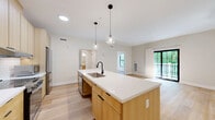 This thoughtfully designed single-floor residence offers a generous layout spread across one level, seamlessly blending modern aesthetics with functional living. The open-concept kitchen and living room serves as the heart of the home, anchored by a sleek kitchen island with light wood cabinetry, white quartz countertops, a classic subway tile backsplash, stainless steel appliances, and pendant lighting overhead. Light-toned hardwood flooring flows consistently throughout the entire space, enhancing the bright, airy atmosphere. The living area opens directly onto a private balcony through sliding glass doors framed in matte black, offering tranquil views of lush, mature greenery. The bedroom is well-proportioned, featuring large black-framed windows that flood the room with natural light and provide views of the surrounding neighborhood. A sliding barn-style door adds a contemporary architectural touch to the bedroom entry. The bathroom is designed with accessibility in mind, featuring a walk-in roll-in shower with neutral tile surround, grab bars, a floating wood vanity with a white countertop, and a well-lit mirror. Additional spaces include a dedicated laundry room, a hallway, and an entrance area, all contributing to a complete and practical floor plan.
