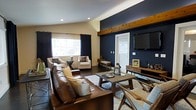 This single-floor space showcases a well-appointed living area that blends contemporary design with warm, rustic accents. The room is anchored by a striking navy blue accent wall that creates a bold focal point, complemented by neutral beige tones on the surrounding walls for a balanced, cohesive palette. A natural wood beam runs horizontally across the upper portion of the navy wall, adding architectural character and a touch of warmth to the space.

The living area features dark hardwood flooring that grounds the room and contrasts elegantly with the lighter furnishings. Seating includes a rich brown leather sofa paired with cream upholstered chairs, offering a layered and inviting arrangement around a rectangular wood coffee table. A soft area rug defines the seating zone and adds texture underfoot.

Natural light enters through a large window dressed with dark curtain panels, providing both brightness and privacy. A wall-mounted flat-screen television is positioned on the navy accent wall above a low-profile wood media console. Vaulted ceiling lines, recessed lighting, and thoughtful décor details complete the polished, functional ambiance of this living space.