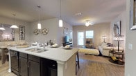 This single-story space showcases an open-concept layout that effortlessly connects the kitchen and living area, creating a bright and functional environment. The kitchen is anchored by a spacious island with granite-style countertops, a double-basin sink, and dark cabinetry that provides a stylish contrast to the light-toned surfaces. Two sleek pendant lights hang above the island, adding focused illumination and a contemporary design touch. The kitchen flows naturally into the living area, which features warm hardwood-style flooring that extends throughout the space. A ceiling fan in the living area contributes to both comfort and airflow. Neutral-toned walls throughout create a clean, versatile backdrop that complements the overall aesthetic. Natural light enters through a set of windows and a glass-paneled door at the far end of the living area, offering views to the exterior and enhancing the sense of openness. Decorative wall accents add personality to the space. The cohesive design and open floor plan make this a welcoming and practical living environment suited for both everyday living and entertaining.