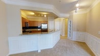 This single-floor residence showcases a thoughtfully designed open-concept layout that seamlessly connects the kitchen and living areas. The kitchen is a standout feature, equipped with stainless steel appliances, including a refrigerator and microwave, complemented by warm wood cabinetry and a classic dark tile backsplash. Granite countertops extend across the kitchen and wrap around a generously sized curved peninsula with elegant white panelled skirting, providing both additional workspace and a natural division between the kitchen and the carpeted living area. Crown moulding runs throughout the space, adding a refined architectural detail that elevates the overall aesthetic. Recessed track lighting illuminates the kitchen, while additional ceiling fixtures contribute to a well-lit and welcoming ambiance. A built-in shelving unit adds a practical yet decorative touch to the living area, ideal for displaying books or décor. Multiple interior doors lead to additional rooms, suggesting a functional floor plan with well-defined private spaces. The neutral colour palette of soft beige tones throughout the carpeting, walls, and trim creates a cohesive and inviting atmosphere suited to a variety of interior styles.
