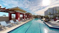 This rooftop pool deck offers a resort-style retreat elevated above the surrounding cityscape. The centerpiece is a generously sized lap pool with crystal-clear turquoise water, its clean geometric lines extending the full length of the deck. A row of modern white lounge chairs lines the poolside, providing ample space for sunbathing and relaxation. A striking covered cabana structure anchors one side of the deck, featuring bold red columns and a tiled roof that lends a Mediterranean-inspired architectural character to the space. The cabana provides welcome shade while maintaining an open, airy feel. The surrounding deck is finished in light-toned pavers that complement the bright, sun-drenched atmosphere. Lush greenery and manicured shrubs accent the perimeter, adding a natural touch to the urban setting. The elevated position of the deck affords panoramic views of the surrounding buildings and open sky, creating a sense of openness and escape. Safety amenities, including a life ring station, are discreetly integrated into the design. Whether used for active lap swimming or leisurely lounging, this rooftop pool deck delivers a polished and inviting outdoor amenity experience.