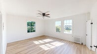 This single-floor unit is designed with a clean, minimalist aesthetic throughout. The main living area is generously proportioned, featuring light-toned hardwood flooring that extends uniformly across the space. Multiple white-framed, multi-pane windows line the walls, flooding the room with abundant natural light and offering views of surrounding greenery and the broader neighborhood. A five-blade ceiling fan with an integrated light fixture is centrally mounted, providing both ventilation and overhead illumination. A classic cast-iron radiator is positioned beneath one of the windows, indicating a traditional heating system.

The kitchen area includes a stainless steel refrigerator set against crisp white walls, maintaining the unit's cohesive, neutral color palette. Additional rooms continue the light hardwood flooring and white wall treatment, creating a consistent, airy feel throughout.

The bathroom is a notable feature, finished with full white subway tile in the tub and shower surround, paired with striking matte black fixtures including the showerhead, faucet, and tub spout. A window within the bathroom provides natural ventilation. The 3D floor plan indicates a straightforward, efficiently organized layout with clearly defined living, sleeping, kitchen, and bathroom areas interconnected by open doorways.