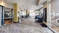 This spacious fitness center is thoughtfully designed to accommodate a wide range of workout styles and fitness goals. The layout is open and expansive, with distinct zones dedicated to cardio, strength training, and functional fitness. A patterned carpet with a hexagonal motif covers much of the floor, while rubber mat sections provide appropriate surfaces beneath weight equipment.

The cardio zone features stair climbers, elliptical trainers, and a stationary bike, all positioned near large floor-to-ceiling windows that flood the space with natural light and offer views of the surrounding outdoor area. Ceiling fans are distributed throughout to maintain airflow and comfort during workouts.

The strength training area is well-appointed with a variety of cable and resistance machines, an adjustable bench, and a full rack of dumbbells displayed along a mirrored wall. The mirrors effectively expand the sense of space while allowing users to monitor their form.

A dedicated functional training zone includes stability balls, medicine balls, a cable station, and a large wall-mounted display screen. Decorative accent walls featuring geometric patterns in olive green and gold add visual interest throughout the space. A water fountain and storage areas near the entrance ensure convenience. Large glass doors provide a welcoming, light-filled entry point.