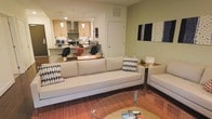 This single-floor residence offers a thoughtfully designed open-concept layout that seamlessly connects the living and kitchen areas, creating a cohesive and functional living environment. The living area is anchored by a generously sized, neutral-toned sofa accented with patterned throw pillows, complemented by a round glass coffee table and a warm terracotta-hued area rug that adds texture and color to the space. Decorative wall art in navy and white tones adds a personalized touch to the room. The kitchen, visible from the living area, features light wood cabinetry, a stainless steel range hood, and pendant lighting that casts a warm glow over the workspace. A kitchen island or counter bar with seating provides an ideal spot for casual dining or working. Recessed lighting throughout ensures the space is well-lit, while the overall color palette of soft greens, warm neutrals, and natural wood tones contributes to a calm and welcoming ambiance. The open flow between areas makes the space feel airy and versatile, well-suited for both relaxing and entertaining.