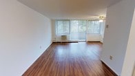 Located at 22701 Lakeshore Blvd in Euclid, Ohio, this single-floor unit offers a practical and well-organized layout. The living area is a standout feature, showcasing rich hardwood-style flooring that runs throughout the space and reflects natural light beautifully. Large windows paired with a sliding glass door line the exterior wall, inviting abundant natural light and providing views of the surrounding greenery. Vertical blinds offer privacy and light control. A ceiling fan adds to the comfort of the living area, while a wall-mounted air conditioning unit and baseboard heating ensure year-round climate control. The open feel of the living space is enhanced by clean white walls that create a bright, neutral backdrop. The kitchen is positioned toward the rear of the unit and features cabinetry providing ample storage, with a layout designed for efficient use of space. A separate entry door is visible from the kitchen area, offering convenient access. The floor plan, as seen in the dollhouse view, flows logically from the living and dining areas through to the kitchen, making the overall layout easy to navigate and well-suited for everyday living.