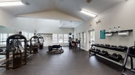 This single-level fitness center offers a spacious and well-appointed workout environment designed to support a wide range of training styles. The room is defined by its striking vaulted ceiling, which creates an open, airy atmosphere and contributes to the generous sense of volume throughout the space. Large windows with white blinds line the walls, allowing ample natural light to fill the interior while providing views of the surrounding greenery.

The flooring consists of durable rubber gym matting, providing both safety and comfort underfoot. The facility is divided into distinct functional zones. The strength training area houses multiple cable and resistance machines, a Smith machine with bench press, and additional selectorized equipment arranged to allow easy movement between stations. A wall-mounted flat-screen television is positioned above the Smith machine, keeping members entertained during workouts.

The functional training zone features a large tire sled, battle ropes, and a full rack of resistance bands mounted along the wall, catering to dynamic and conditioning-based exercise. A well-stocked dumbbell rack lines one wall, offering a broad range of free weights. Mirrored walls in select areas enhance the sense of space. Ceiling fans and fluorescent lighting ensure comfort and visibility throughout. The facility is bright, clean, and comprehensively outfitted for a full-body training experience.