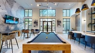 Located at 6851 N 93rd Ave in Glendale, Arizona, this contemporary community clubroom is a well-appointed social and recreation space designed for both leisure and entertainment. Soaring double-height ceilings create a dramatic sense of openness, while an expansive wall of floor-to-ceiling black-framed windows and glass doors floods the interior with natural light and offers unobstructed views of the surrounding palm tree-lined courtyard.

The centerpiece of the room is a solid wood billiards table set atop a patterned area rug, providing a focal point for social gatherings. A large-scale abstract stone mural dominates one wall, adding an artistic, textural element that anchors the space. Opposite, a stylish wet bar with a white stone countertop, dark cabinetry, open metal shelving, and pendant lighting with gold-accented shades serves as a sleek gathering hub with bar-height seating.

A cozy arched nook with upholstered bench seating and a bistro table offers an intimate retreat within the larger room. Additional seating areas are distributed throughout, including bar-height tables and mixed accent chairs. Light-toned large-format tile flooring flows seamlessly across the entire space, complementing the bright, modern aesthetic. The overall ambiance is sophisticated yet inviting, ideal for community socializing and recreation.