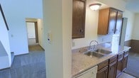 This two-story residence offers a practical and comfortable layout designed for everyday living. The kitchen is a central feature, showcasing granite-style speckled countertops paired with warm, medium-toned wood cabinetry that provides ample upper and lower storage. A stainless steel double-basin sink is positioned beneath overhead cabinetry, complemented by a classic chrome faucet and flush-mount ceiling lighting that keeps the space well-lit. The kitchen's layout includes a structural column that partially separates it from the adjoining living areas, creating a natural transition between spaces. Beyond the kitchen, the residence opens into carpeted hallway and living areas, visible through the passageway, suggesting a connected and flowing floor plan. The neutral wall tones throughout contribute to a clean, understated aesthetic that allows for versatile furnishing options. The two-floor configuration adds spatial dimension to the residence, offering separation between living and private areas. Overall, the space presents a straightforward and functional interior with classic finishes suited to comfortable day-to-day living.