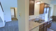 This two-story residence offers a practical and comfortable layout designed for everyday living. The kitchen is a central feature, showcasing granite-style speckled countertops paired with warm, medium-toned wood cabinetry that provides ample upper and lower storage. A stainless steel double-basin sink is positioned beneath overhead cabinetry, complemented by a classic chrome faucet and flush-mount ceiling lighting that keeps the space well-lit. The kitchen's layout includes a structural column that partially separates it from the adjoining living areas, creating a natural transition between spaces. Beyond the kitchen, the residence opens into carpeted hallway and living areas, visible through the passageway, suggesting a connected and flowing floor plan. The neutral wall tones throughout contribute to a clean, understated aesthetic that allows for versatile furnishing options. The two-floor configuration adds spatial dimension to the residence, offering separation between living and private areas. Overall, the space presents a straightforward and functional interior with classic finishes suited to comfortable day-to-day living.
