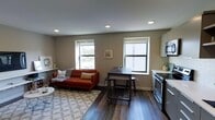 This single-floor space showcases a thoughtfully designed open-concept layout that seamlessly integrates the living and kitchen areas into one cohesive environment. Dark hardwood-style flooring runs throughout, providing a warm contrast to the neutral gray walls and white ceiling with recessed lighting. The living area is anchored by a rust-orange sofa set over a patterned area rug, creating a cozy yet functional seating zone. Two double-hung windows on the main wall allow generous natural light to fill the space, offering views of the surrounding outdoors. A compact dining area with a dark wood table and industrial-style stools serves as a natural divider between the living and kitchen zones. The kitchen, positioned along one wall, features stainless steel appliances — including a range and over-the-range microwave — white cabinetry, and light countertops, delivering a clean, modern aesthetic. The overall design is efficient and practical, maximizing the use of available space while maintaining a comfortable, welcoming ambiance suited to everyday living.