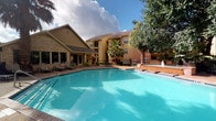 This outdoor pool area offers a well-appointed recreational retreat designed for relaxation and leisure. The centerpiece is a generously sized swimming pool with clear, turquoise water, accessible via stainless steel handrails and steps. Adjacent to the main pool sits a raised hot tub or spa feature, complete with a decorative fountain that adds both visual appeal and a soothing ambient sound to the environment.

The pool deck is finished in a light-toned concrete surface that surrounds the water on multiple sides, providing ample space for lounge chairs and patio seating. Numerous chaise lounges and chairs are arranged along the deck, offering plenty of options for sunbathing or casual gathering. Patio umbrellas are stationed throughout the area, providing shade during sunny days.

Mature trees, including large shade trees and tall palm trees, frame the perimeter of the pool area, creating a lush, park-like atmosphere. A low wrought iron fence encloses the space, ensuring privacy and security. A dedicated pool house or amenity building with wood-framed glass doors provides convenient access to the pool deck, with posted pool rules and exterior wall-mounted lighting visible near the entrance. The overall ambiance is open, bright, and inviting.