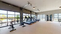 This single-floor fitness center is thoughtfully designed to support a comprehensive workout experience. The space is divided into distinct training zones, each tailored to a specific type of exercise. The cardio area is anchored by a row of commercial-grade treadmills, stationary bikes, and elliptical machines, all positioned in front of large glass garage-style doors that open to outdoor views, creating a bright and airy atmosphere. These oversized doors serve as a standout architectural feature, inviting abundant natural light and offering visual connection to the surrounding outdoor amenity areas, including what appears to be a pool terrace with lush plantings. A dedicated functional training zone houses TRX suspension trainers, rowing machines, and spin bikes, also benefiting from glass door access to the outdoors. The strength and free weights area is well-stocked with a full dumbbell rack, cable machines, battle ropes, plyo boxes, medicine balls, and stability balls organized on a tiered rack. A glass-partitioned wall separates portions of the space, adding a sleek, modern design element. Neutral-toned rubber flooring, recessed lighting, and soft gray walls contribute to a clean, professional ambiance throughout the facility.